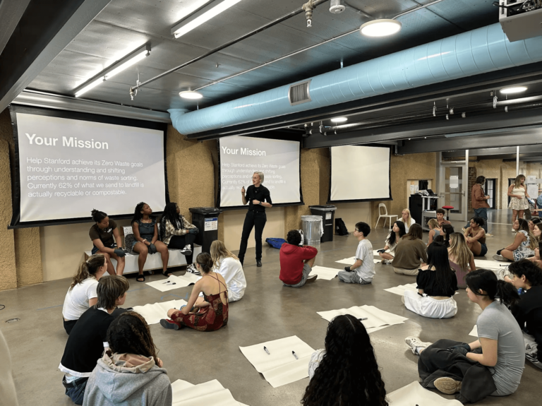 Students grouped in pairs sitting on floor with large piece of paper and sharpie per pair watching presentation
