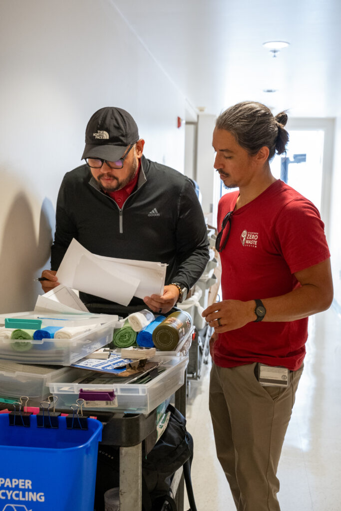 Two people looking at papers with a cart with containers of bags and labels on top.