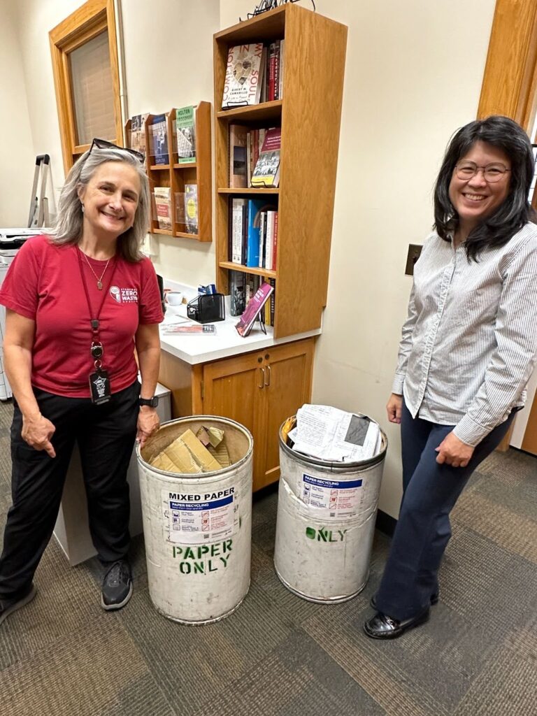 Two people standing with two circular bins that say paper only.