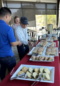 3 people going through the line at a buffet table with sandwiches and drinks in reusable containers