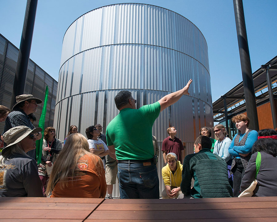 A group of people attentively listening to a guide in front of a large cylindrical metal tank on a sunny day.