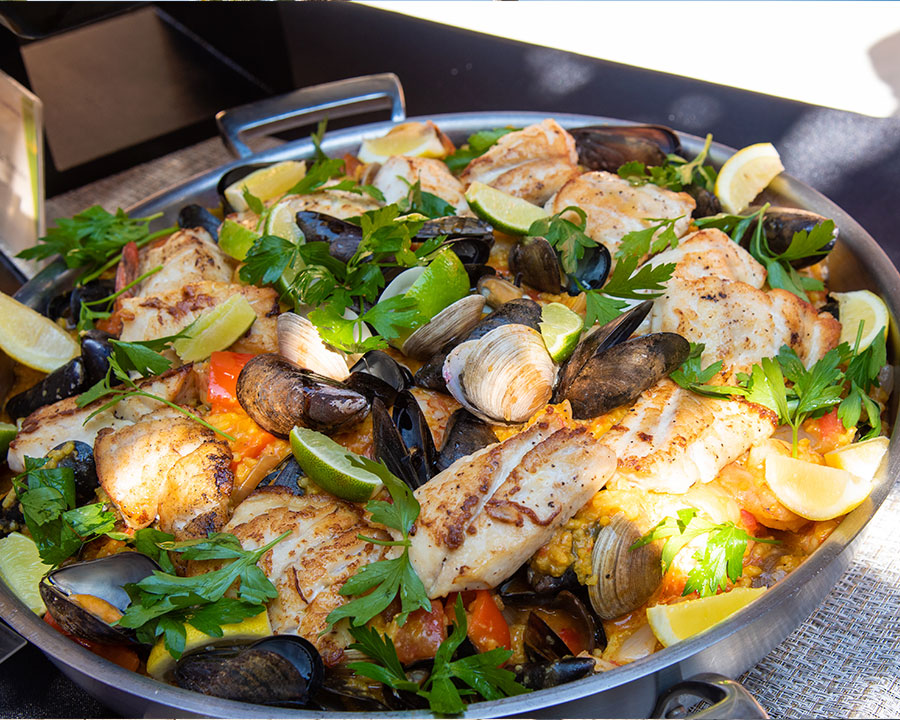 A vibrant seafood paella dish with fish, mussels, clams, lemon wedges, and fresh parsley in a serving pan.