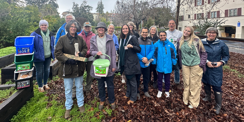 A group of staff and faculty posing for a picture on a rainy day with recycle, compost, and landfill bins.