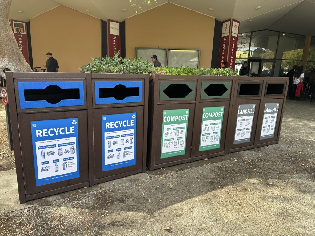 Two recycle, two compost, and two landfill bins together outside a buildings.