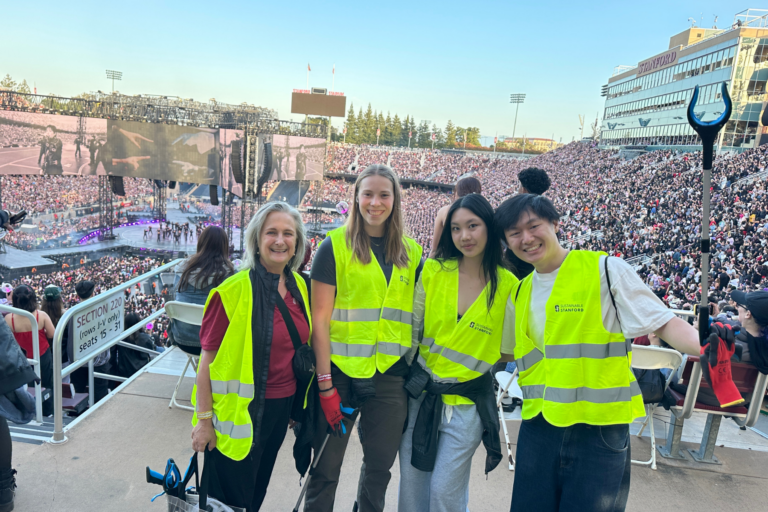 Four people with yellow vests posing with a background of a large crowd in an outdoor stadium with a stage and large screens showing the performers.