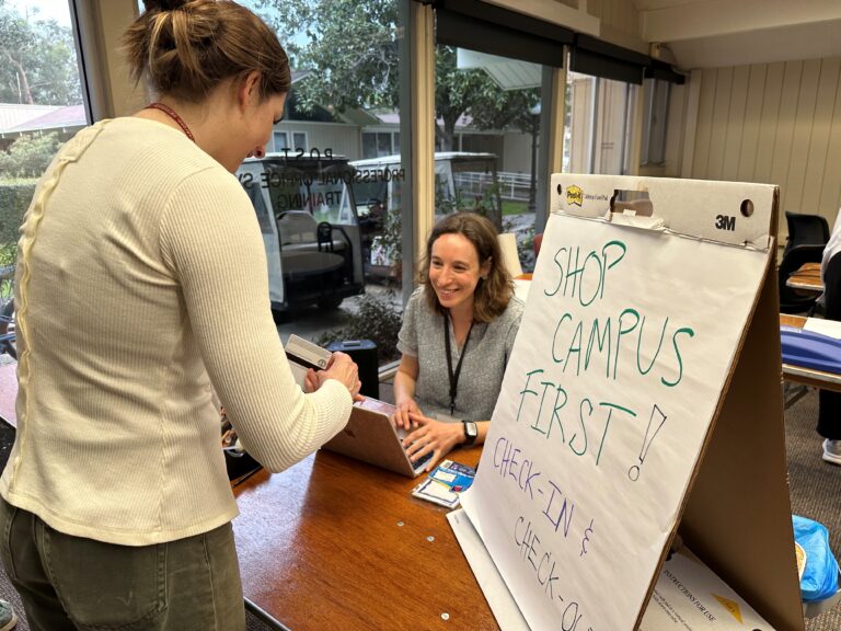 One woman sitting at a check-in table with a laptop checking another woman's ID, smiling next to a sign that says "Shop Campus First"